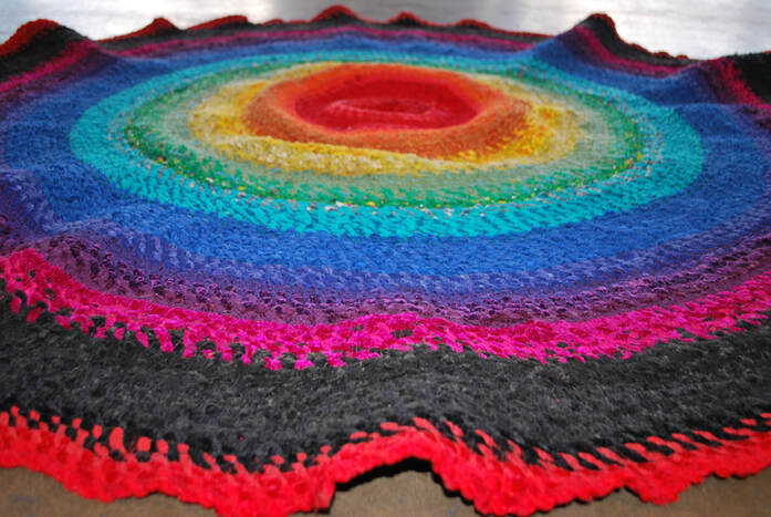 A round rag rug in tiers of rainbow colors: red, orange, yellow, green, blue, purple, acid pink, black, and red.