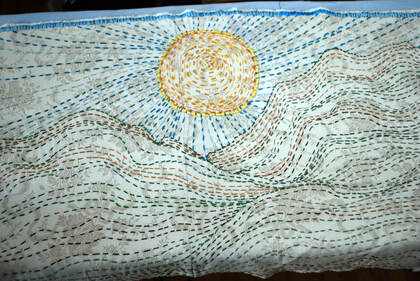 Embroidered panel of green mountains, yellow sun, and blue sky on beige fabric.