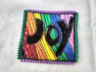 On a three inch by three inch panel, rainbow stripes outline the word JOY. Light purple and green zipper teeth outline the square panel.