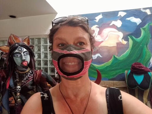 A smiling woman wears a face mask with a clear window over the mouth. In the background is a colorful painting and a blue-faced sculpture with a crown, three eyes, and a red tongue.
