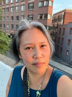 Grey haired Asian woman with a blue top in front of brick apartment buildings