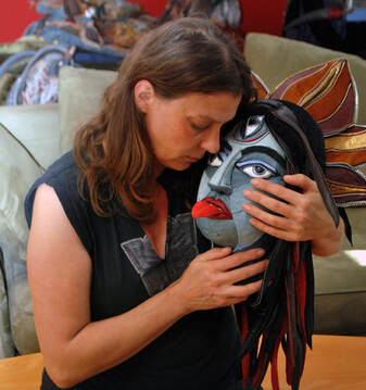 A white woman with light brown hair cradles a sculpture of the goddess Kali's head in her hands. Kali is blue, has three eyes and a tall crown, and sticks out her red tongue.