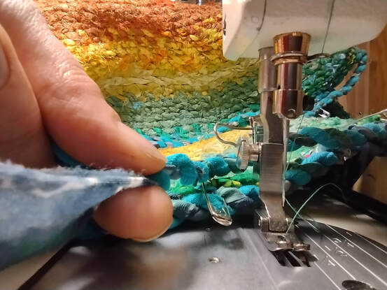 A hand twists turquoise rag twin next to a sewing machine. In the background are rows of rag twine sewn together in different colored tiers: turquoise, green, pale green, yellow, and orange.
