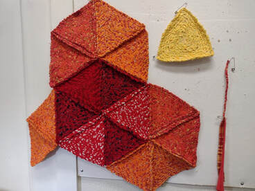 A rag rug made of red and orange triangles with a yellow rag triangle and a twist of orange rag twine hanging nearby.
