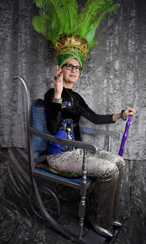A woman in a green and gold feathered headdress sits on a silver and blue stylized wheelchair.