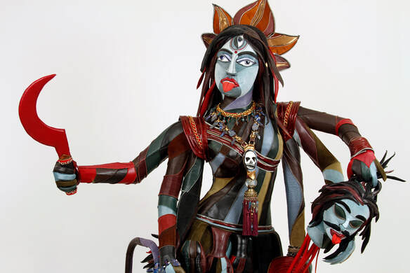 A multi-colored, multi-limbed goodess with three eyes and six arms built into a walker. She holds a red scythe in her upper right hand and a blue demon head in her upper left. 