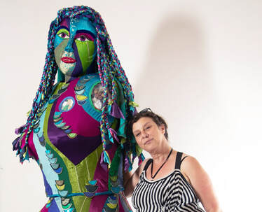 A woman in balck and white stripes with brown hair leans against a large sculpture of a colorful female figure with braided hair  and scales running up and down her body.