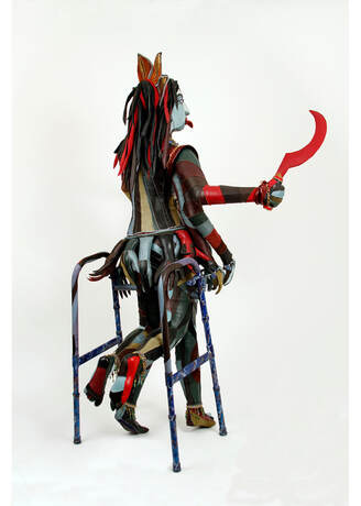 Back view of a sculpture of the goddess Kali built into a walker. The sculpture has multicolored skin and red and black hair. Three right arms are visible, and the top hand holds a red scythe.arms are visible