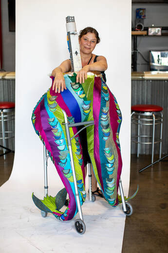 A smiling woman in back of a sculpture of a pair of colorful legs/tail fins built into a walker.