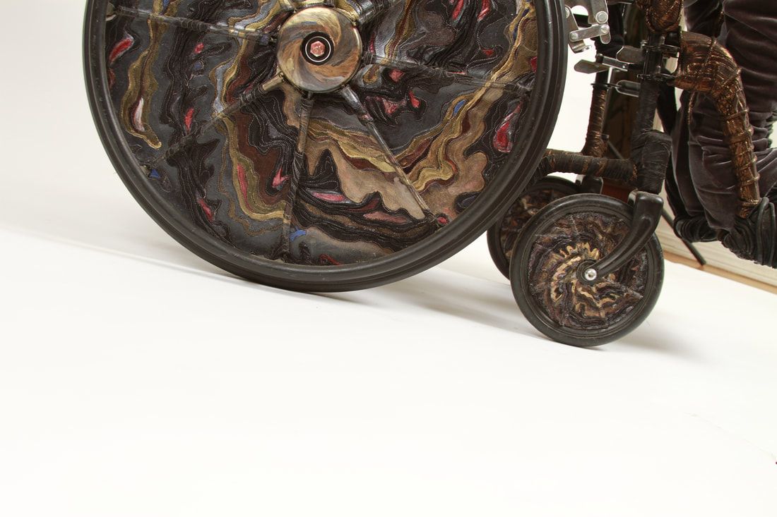 Close up of a manual wheelchair's wheels covered in black, gold, and copper quilted swirls with spots of red and blue.
