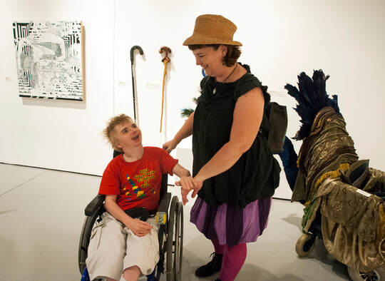 A sandy haired  young man in a wheelchair is on the left, a standing woman in balck with purple striped bloomers and a hat is on the right. In the background is a painting, two canes, and a peacock scooter.