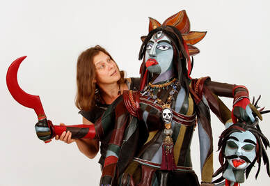 A woman with light brown hair holds the upper arm of a sculpture of the goddess Kali. Kali has multicolored skin, six arms, and three eyes.