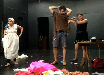 On the left is a woman in a white staple draped dress with a blue ribbon bow. On the right, a woman in black prepares to staple fabric around a man to form a tunic top.
