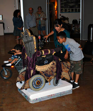 A group of people look closely at a wheelchair that is re-upholstered to look like a throne.