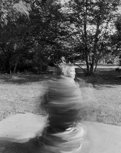 A black and white photo of a whirling bulrred wheelchair with a head faintly visible on top. In the background are grasses and trees.