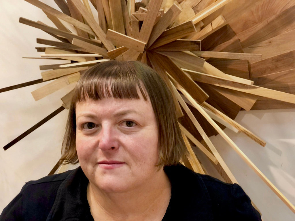A smiling woman with short, light brown hair in bangs in front of a wooden sculpture 