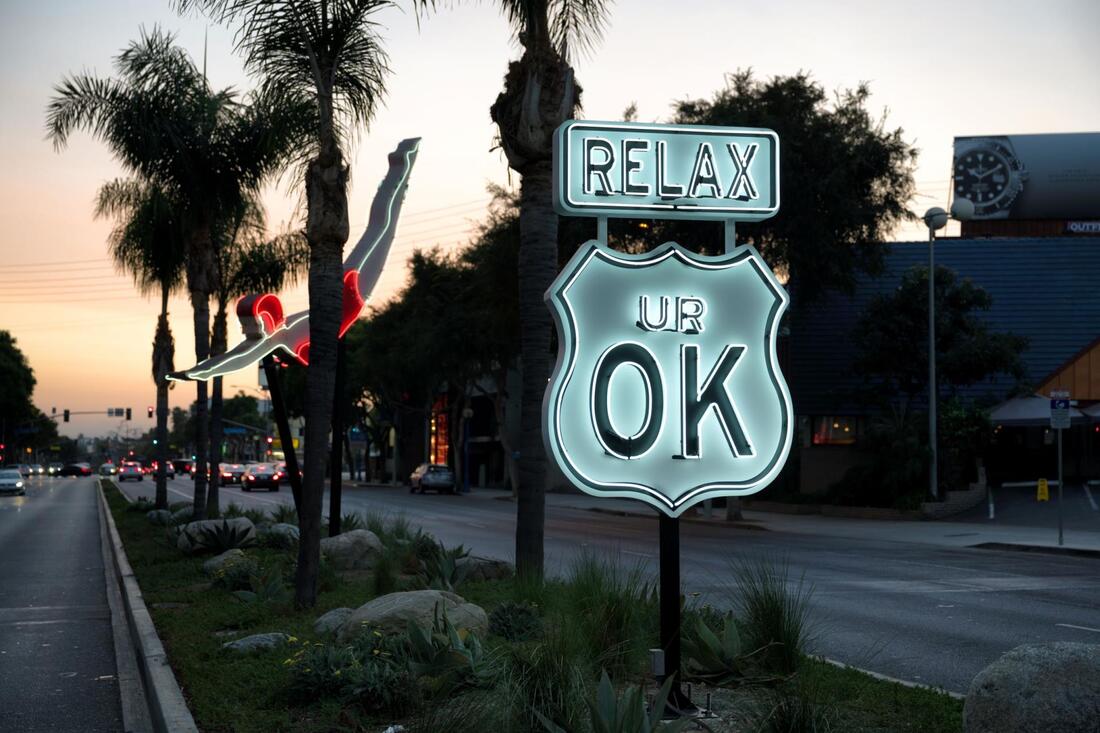 A grassy street median with a neon sign reading RELAX UR OK. Behind it is a neon diving woman in between palm trees.